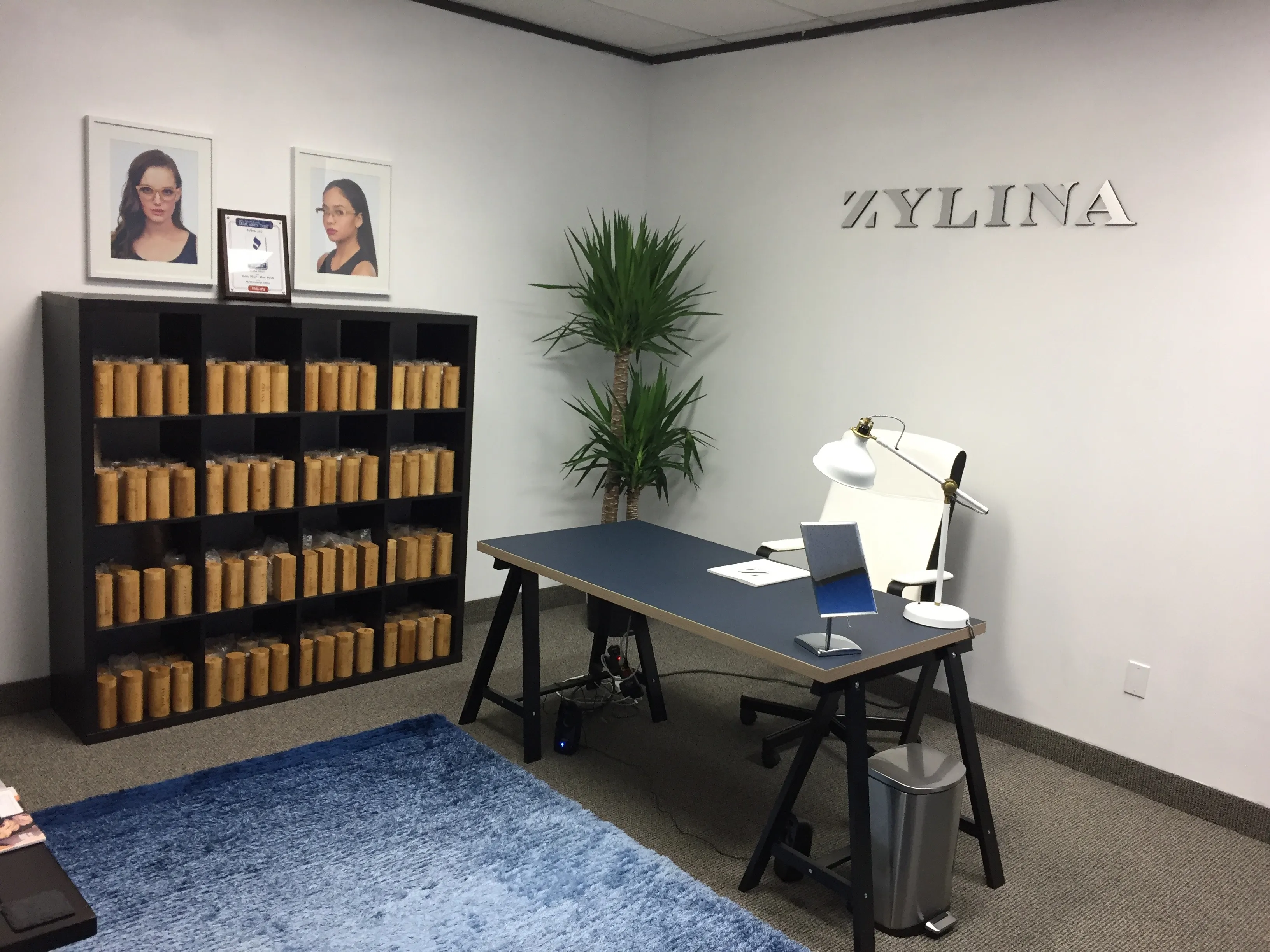 Zylina showroom with ZYLINA letters on the wall, a shelf of bamboo sunglasses tubes, framed model portraits, and a fitting desk with a mirror